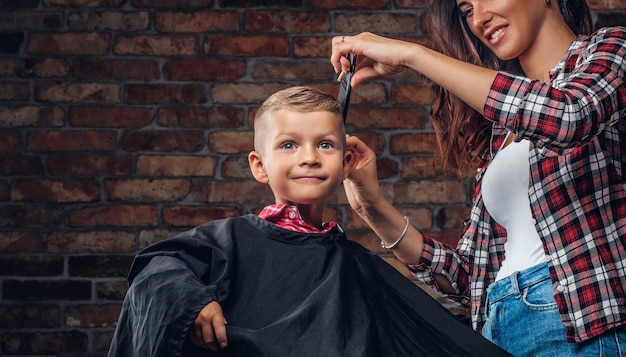 Smiling preschooler boy getting haircut. Children hairdresser with scissors and comb is cutting little boy in the room with loft interior.