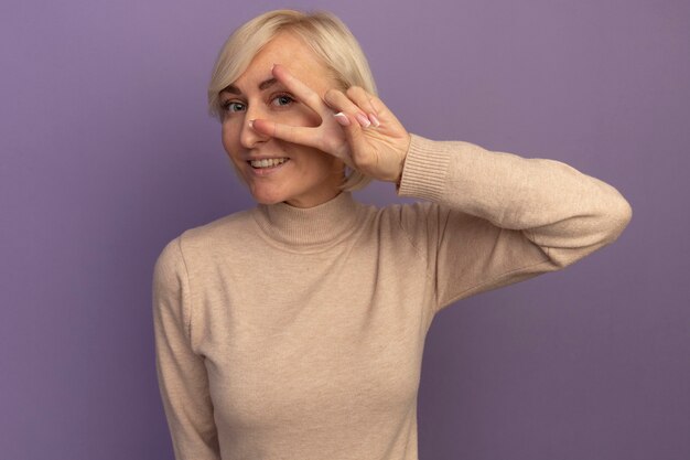 Улыбающаяся симпатичная славянская блондинка жестикулирует знак победы рукой, глядя в камеру сквозь пальцы на фиолетовом
