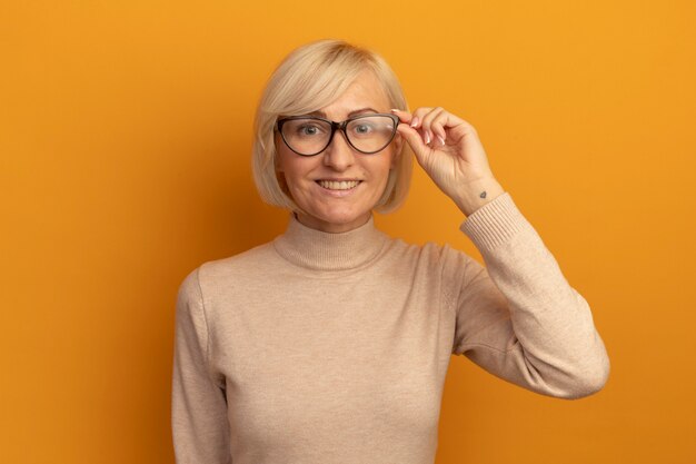 Улыбающаяся симпатичная славянская блондинка смотрит в камеру через оптические очки на оранжевом