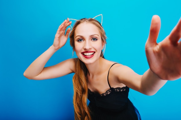 Smiling pretty blonde with blue eyes posing, taking selfie, enjoying party. Wearing black dress and headband with cat ears.