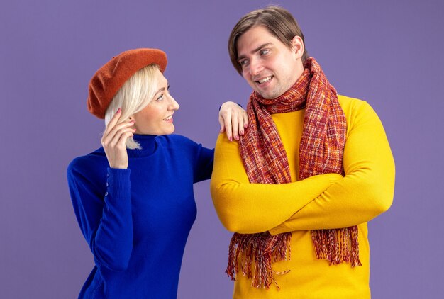 Smiling pretty blonde woman with beret looking at handsome slavic man with scarf around his neck standing with crossed arms isolated on purple wall with copy space