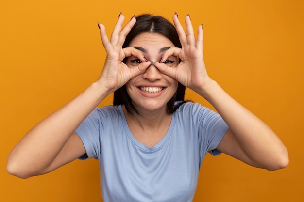 Free photo smiling pretty brunette caucasian girl looks at camera through fingers on orange