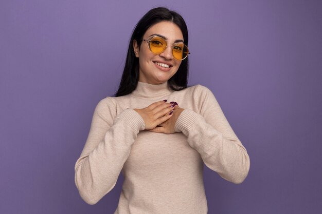 Улыбающаяся симпатичная брюнетка кавказская девушка в солнцезащитных очках кладет руки на грудь на фиолетовом