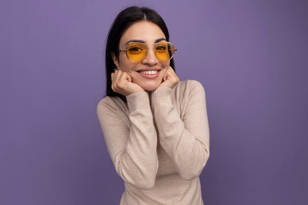 Улыбающаяся симпатичная кавказская девушка брюнетка в солнцезащитных очках кладет руки на подбородок на фиолетовом