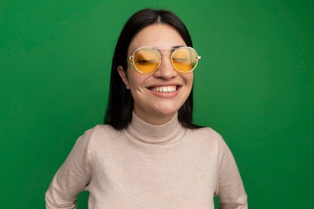 Улыбающаяся красивая брюнетка кавказская девушка в солнцезащитных очках стоит с закрытыми глазами на зеленом