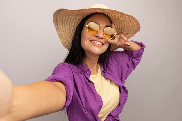 Smiling pretty brunette caucasian girl in sun glasses with beach hat gestures victory hand sign and pretends to hold camera taking selfie on white