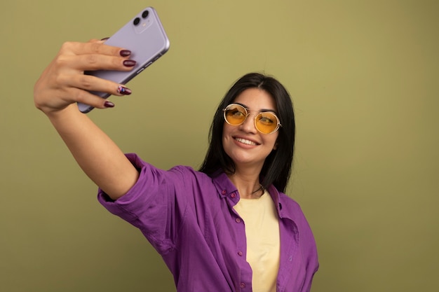 Free photo smiling pretty brunette woman in sun glasses holds and looks at phone taking selfie isolated on olive green wall