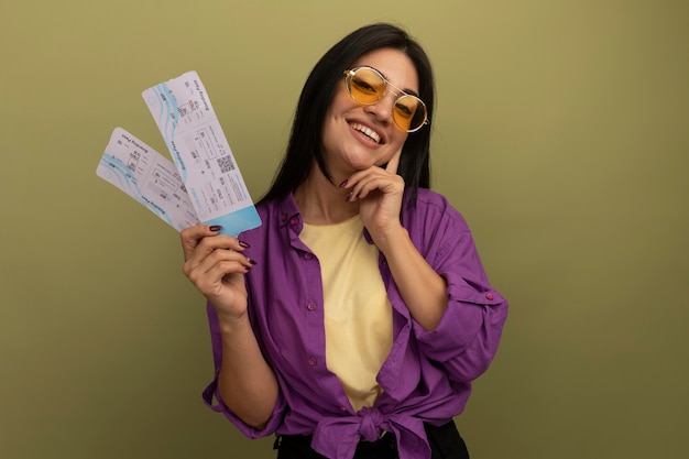 Free photo smiling pretty brunette woman in sun glasses puts hand on face and holds air tickets isolated on olive green wall