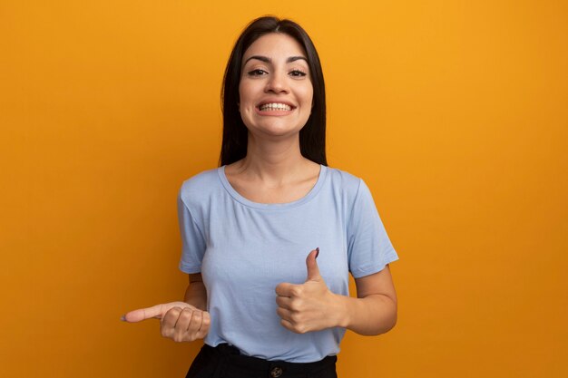 Smiling pretty brunette woman thumbs up and points at side isolated on orange wall