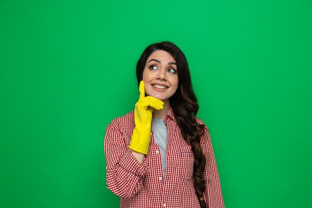 Smiling pretty caucasian cleaner woman with rubber gloves putting hand on her face and looking at side