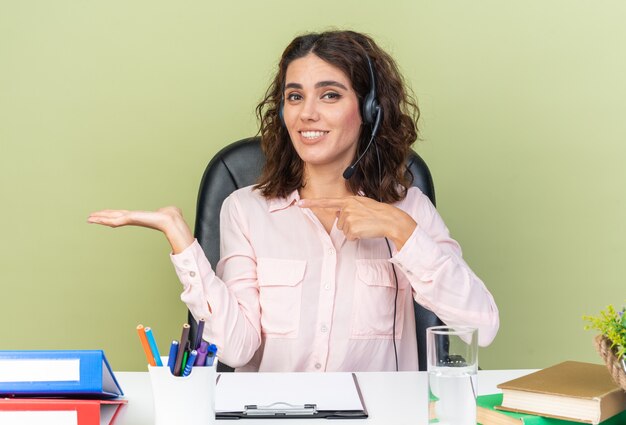 Smiling pretty caucasian female call center operator on headphones sitting at desk with office tools pointing at her empty hand 