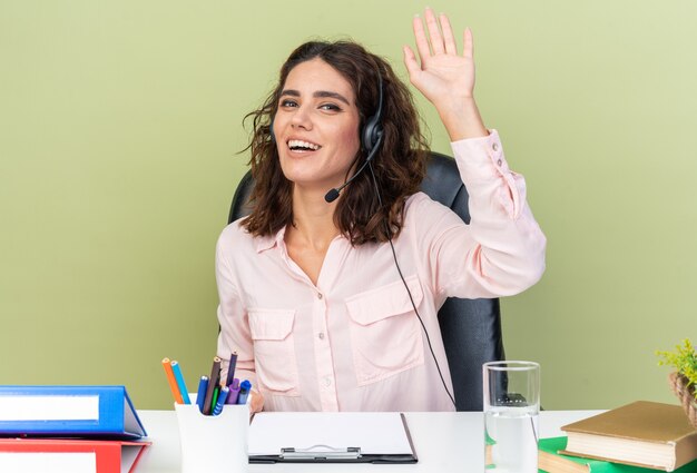 Smiling pretty caucasian female call center operator on headphones sitting at desk with office tools raising hand up 