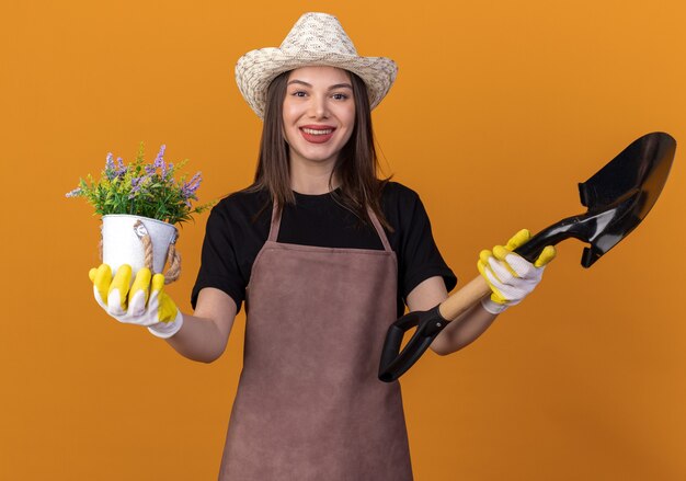 Smiling pretty caucasian female gardener wearing gardening hat and gloves holds flowerpot and spade isolated on orange wall with copy space