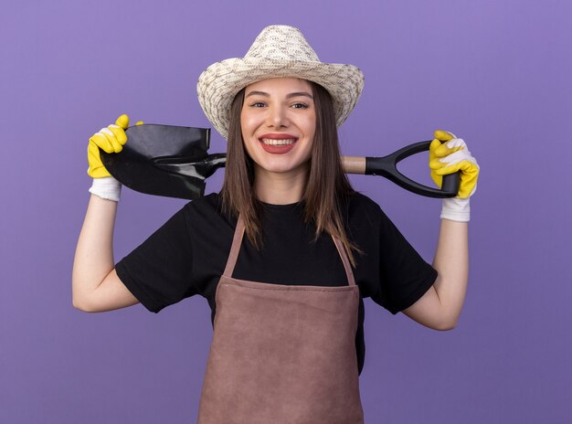 Smiling pretty caucasian female gardener wearing gardening hat and gloves holds spade on neck behind