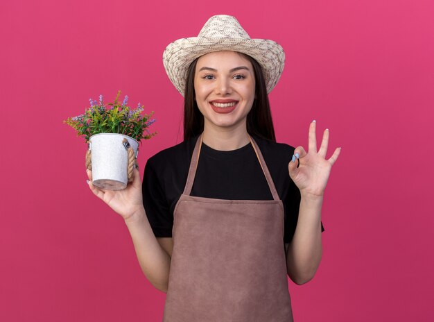 Smiling pretty caucasian female gardener wearing gardening hat holding flowerpot and gesturing ok sign on pink