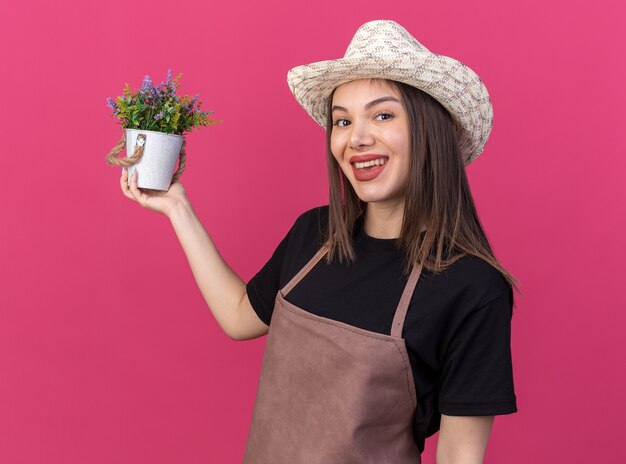 コピースペースとピンクの壁に分離された植木鉢を保持しているガーデニング帽子をかぶってかなり白人女性の庭師の笑顔