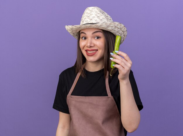 Smiling pretty caucasian female gardener wearing gardening hat holding part of broken hot pepper isolated on purple wall with copy space