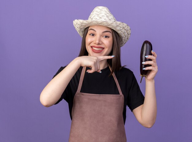 Smiling pretty caucasian female gardener wearing gardening hat holding and pointing at eggplant on purple