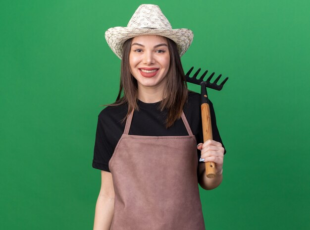 Smiling pretty caucasian female gardener wearing gardening hat holding rake on shoulder isolated on green wall with copy space