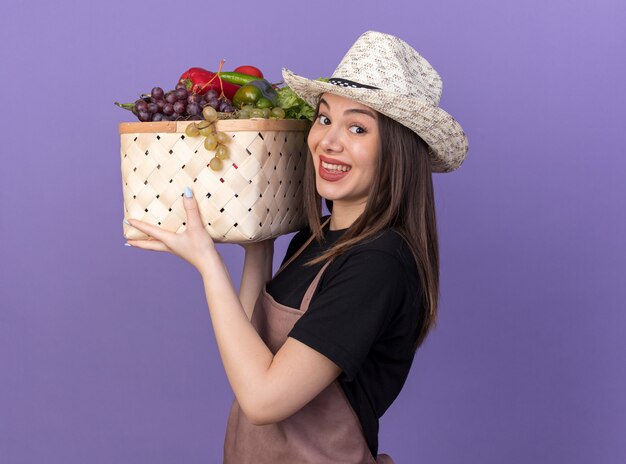 Smiling pretty caucasian female gardener wearing gardening hat holding vegetable basket on purple