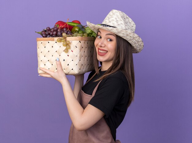紫の野菜バスケットを保持しているガーデニング帽子をかぶってかなり白人女性の庭師の笑顔