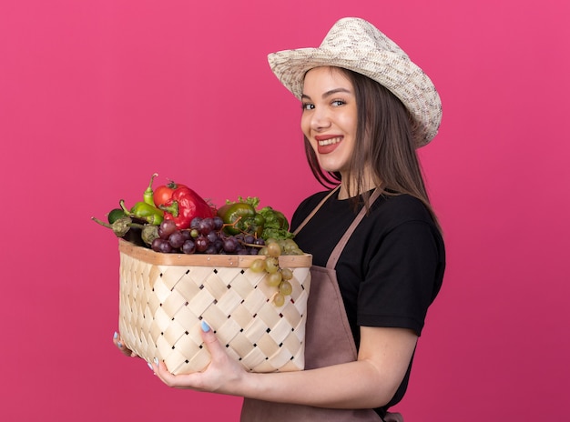 Smiling pretty caucasian female gardener wearing gardening hat stands sideways holding vegetable basket isolated on pink wall with copy space