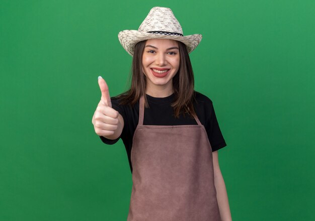 Smiling pretty caucasian female gardener wearing gardening hat thumbs up isolated on green wall with copy space