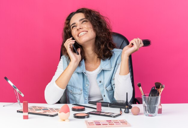 Smiling pretty caucasian woman sitting at table with makeup tools talking on phone and holding comb isolated on pink wall with copy space