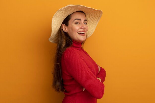 Smiling pretty caucasian woman with beach hat stands sideways with crossed arms on orange