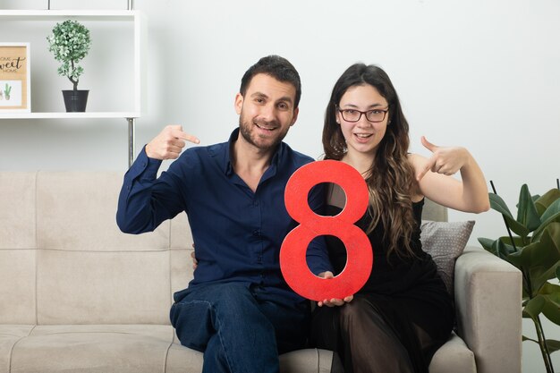 Smiling pretty couple holding and pointing at red eight figure sitting on couch in living room on march international women's day