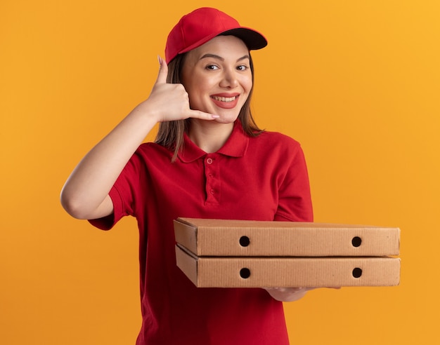 Sorridente bella donna di consegna in gesti uniformi mi chiama segno della mano e tiene scatole di pizza isolate sulla parete arancione con spazio di copia