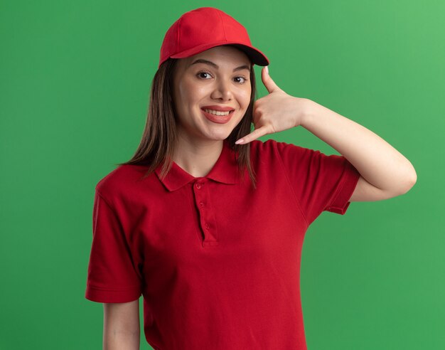 Smiling pretty delivery woman in uniform gestures call me sign isolated on green wall with copy space