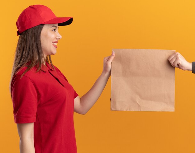 Smiling pretty delivery woman in uniform gives paper package to someone isolated on orange wall with copy space