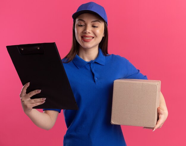 Улыбающаяся красивая женщина-доставщик в униформе держит картонную коробку и смотрит на буфер обмена, изолированный на розовой стене с копией пространства