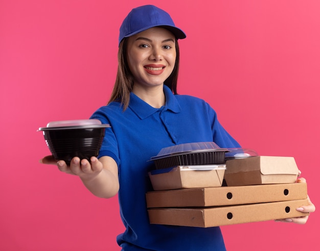 Улыбающаяся красивая женщина-доставщик в униформе держит контейнер для еды и продуктовые пакеты на коробках для пиццы