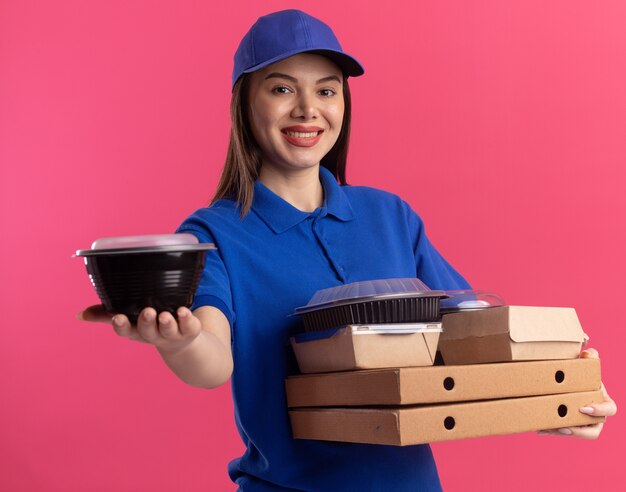 Smiling pretty delivery woman in uniform holds food container and food packages on pizza boxes