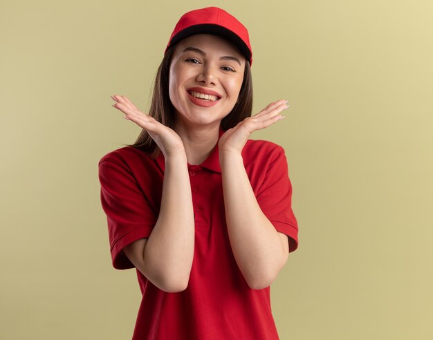 Smiling pretty delivery woman in uniform holds hands close to face isolated on olive green wall with copy space