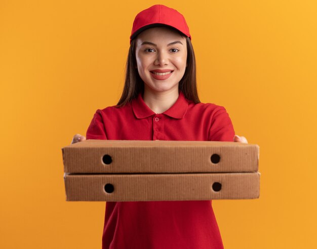 Smiling pretty delivery woman in uniform holds out pizza boxes isolated on orange wall with copy space