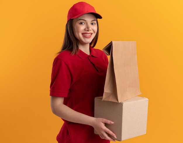 Smiling pretty delivery woman in uniform holds paper package on cardbox isolated on orange wall with copy space