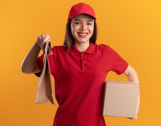 Smiling pretty delivery woman in uniform holds paper package and cardbox on orange