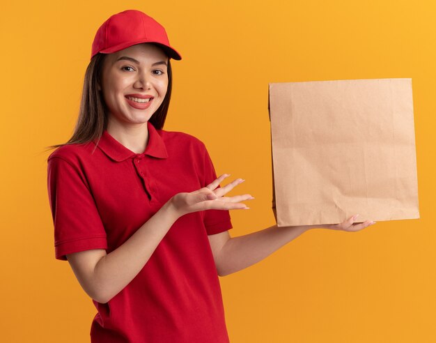 Smiling pretty delivery woman in uniform holds and points at paper package on orange