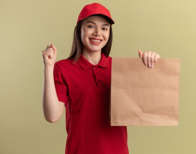 Smiling pretty delivery woman in uniform keeps fist and holds paper package on olive green