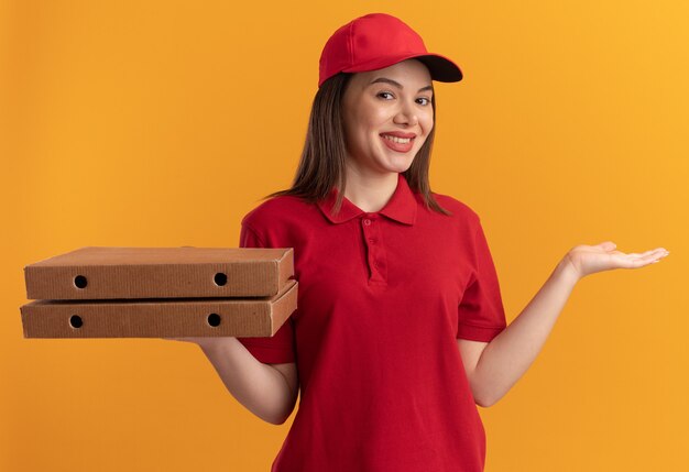 Smiling pretty delivery woman in uniform keeps hand open and holds pizza boxes isolated on orange wall with copy space