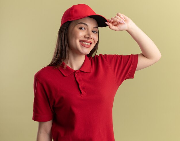制服を着た笑顔のかわいい配達の女性は、コピースペースでオリーブグリーンの壁に分離されたキャップに手を置きます