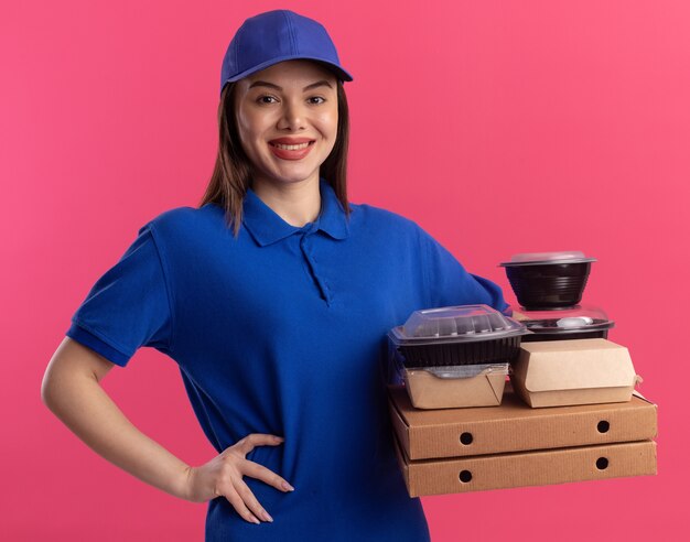 Smiling pretty delivery woman in uniform puts hand on waist and holds food package and containers on pizza boxes isolated on pink wall with copy space