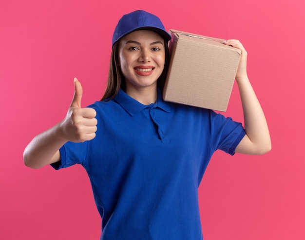 Smiling pretty delivery woman in uniform thumbs up and holds cardbox on shoulder