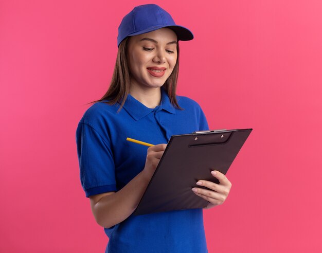 Smiling pretty delivery woman in uniform writes on clipboard with pencil