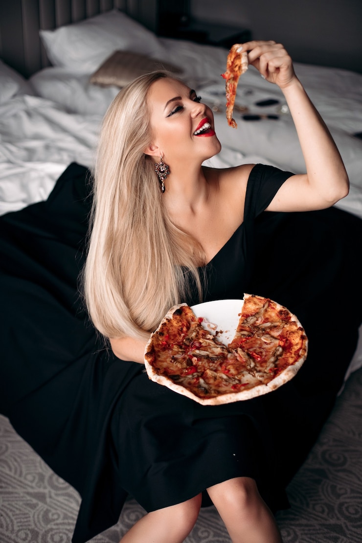 Free Photo smiling pretty fashionable woman in black dress holding a piece of pizza while sitting on the bed in her apartment