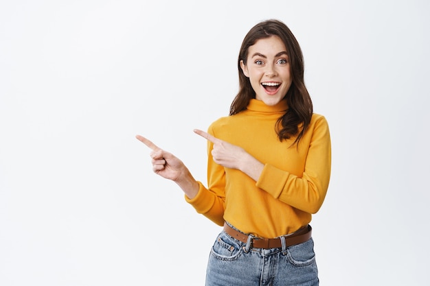 Smiling pretty female model pointing fingers left at empty space and inviting to check it out, showing advertisement on white wall