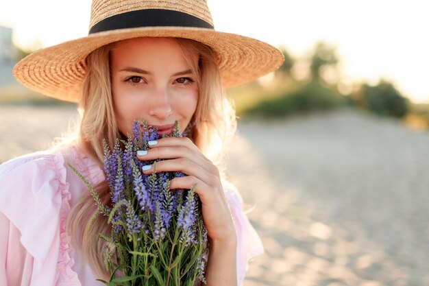 Улыбающаяся красивая девушка в соломенной шляпе позирует на солнечном пляже возле океана с букетом цветов. Крупным планом портрет.